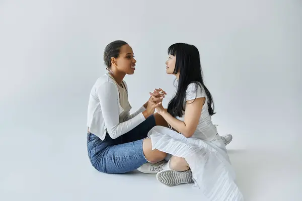 Two young women, one Black and one Asian, sit facing each other on a white background, holding hands. — Stock Photo
