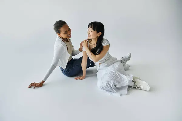 Zwei junge Frauen, eine Afroamerikanerin und eine Asiatin, sitzen auf weißem Hintergrund und teilen einen zärtlichen Moment. — Stockfoto