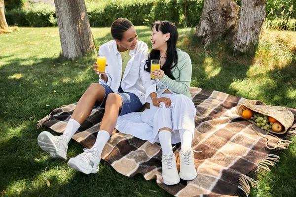 Due giovani donne, una afroamericana e una asiatica, si godono un pranzo al sacco e condividono una risata all'ombra degli alberi. — Foto stock