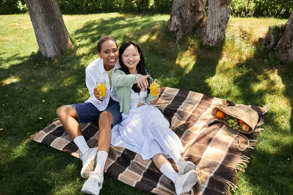 Two young women, one African American and one Asian, enjoy a sunny picnic in a lush park. — Stock Photo