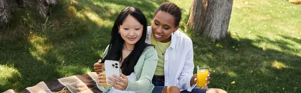 Um casal lésbico multicultural desfruta de um piquenique juntos em um parque, compartilhando uma risada enquanto olha para seu telefone. — Fotografia de Stock