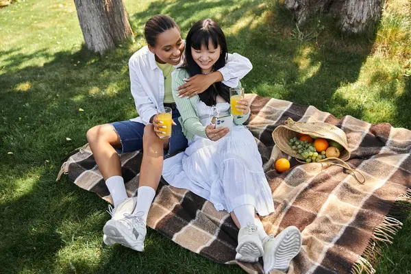 A young lesbian couple enjoys a sunny picnic in the park, embracing each other and sharing a refreshing drink. — Stock Photo