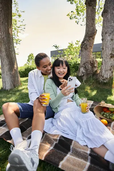 A lesbian couple enjoys a picnic together on a sunny day, sharing a laugh as they look at a phone. — Stock Photo