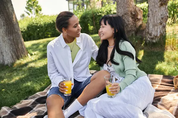Two young women, one African American and one Asian, enjoy a picnic in the park, sharing a smile and drinks. — Stock Photo