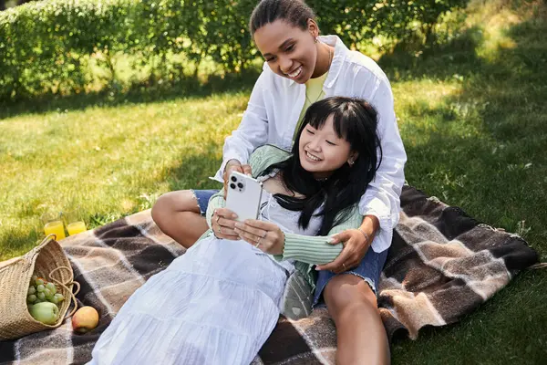 Zwei Frauen genießen ein entspanntes Picknick im Park, während sie gemeinsam auf ein Telefon schauen. — Stockfoto