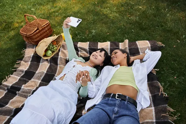 A young lesbian couple enjoys a picnic in the park, taking a selfie with a smartphone. — Stock Photo
