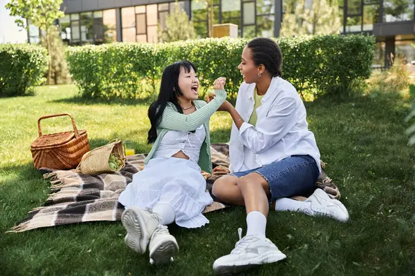 Una pareja lesbiana multicultural disfruta de un picnic en un día soleado, riendo y compartiendo comida en una manta extendida sobre la hierba verde. - foto de stock