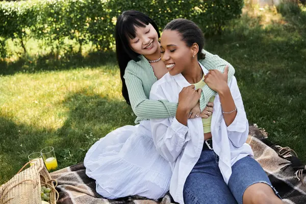 Two women, one African American and one Asian, share a tender embrace during a picnic in a grassy park, enjoying a sunny afternoon. — Stock Photo