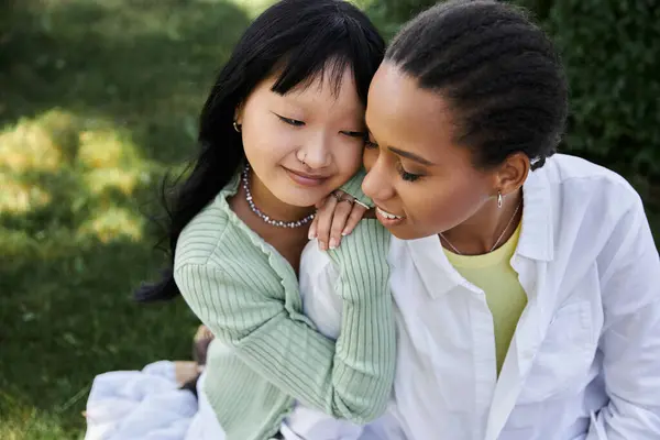 Two young women, one Asian and one African American, share a tender moment while enjoying a picnic in the park. — Stock Photo