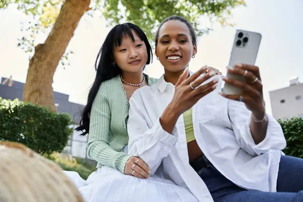 A young lesbian couple enjoys a picnic in the park, sharing a laugh and capturing the moment on their phone. — Stock Photo