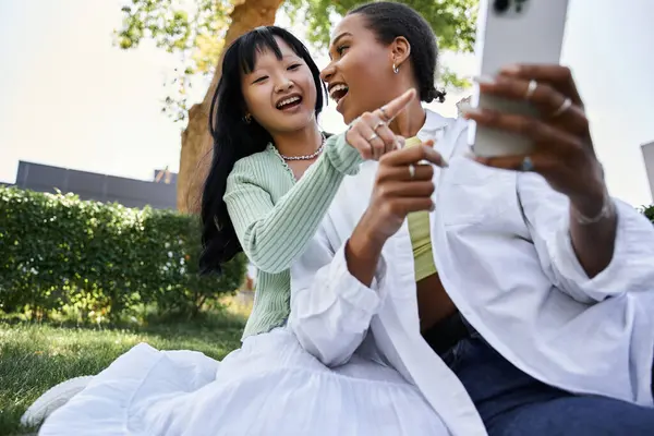 Deux jeunes femmes, une afro-américaine et une asiatique, rient ensemble alors qu'elles sont assises sur une pelouse verte. — Photo de stock