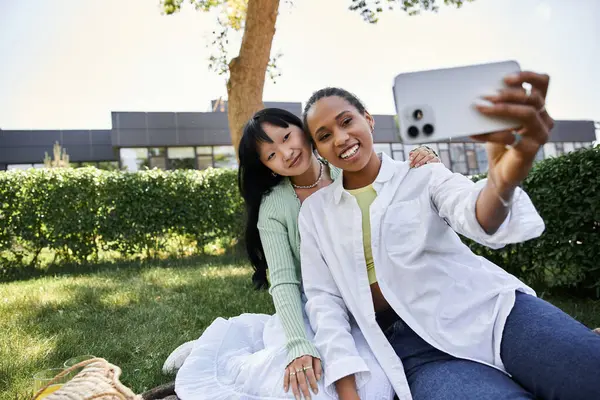 Two young women, one African American and one Asian, are sitting on a blanket in a grassy park, taking a selfie with a smartphone. — Stock Photo