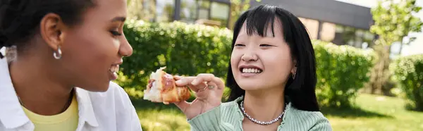 Due giovani donne, una afroamericana e l'altra asiatica, condividono una risata e un boccone da mangiare durante un picnic. — Foto stock