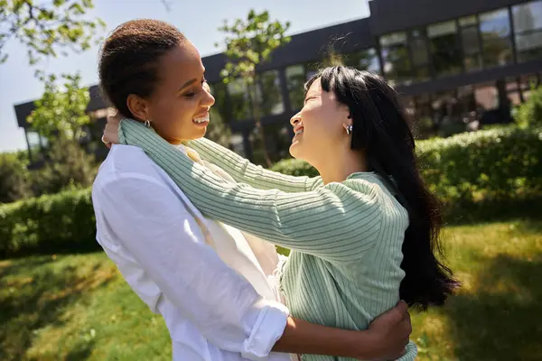 Une embrassade affectueuse entre un couple de lesbiennes noires et asiatiques à l'extérieur, entouré de verdure. — Stock Photo