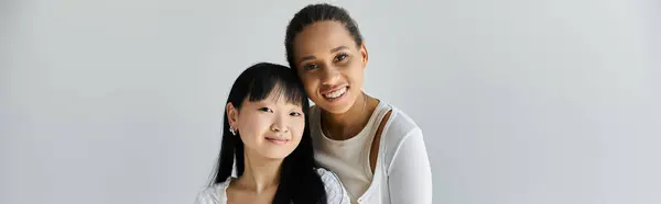 Due giovani donne, una afroamericana e una asiatica, condividono un abbraccio amorevole su un semplice sfondo grigio. — Foto stock