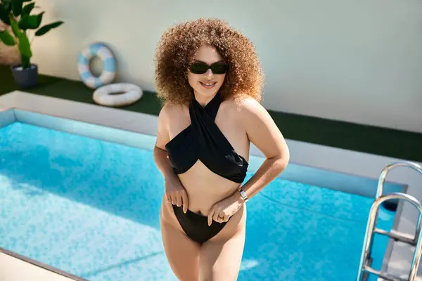 A beautiful woman with curly hair poses in a black swimsuit by the pool on a sunny day. — Stock Photo