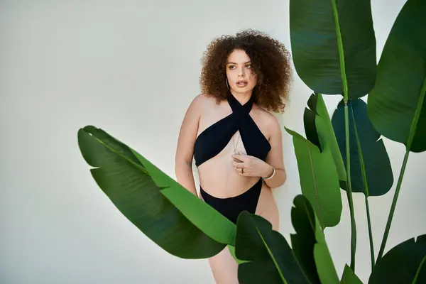 A woman with curly hair, wearing a black swimsuit, poses near a plant on a bright summer day. — Stock Photo