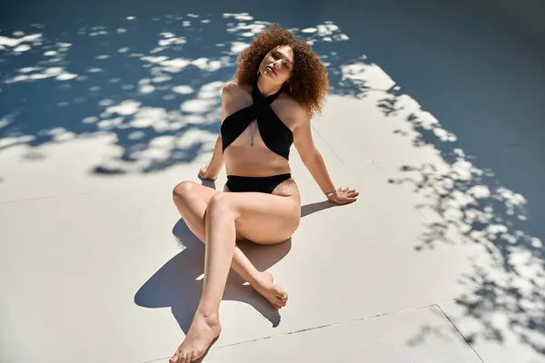 A woman with curly hair in a black swimsuit poses on the edge of a pool on a sunny summer day. — Stock Photo