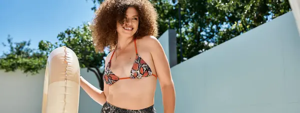 A woman with curly hair, wearing a bikini, smiles as she holds an inflatable pool float. — Stock Photo