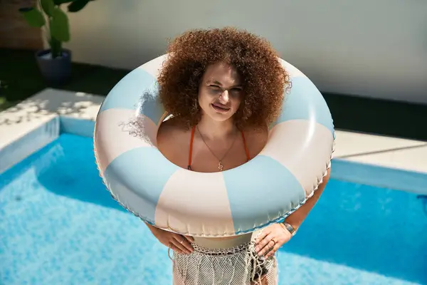 A woman with curly hair in a bikini stands by the pool with a blue and white inflatable ring. — Stock Photo