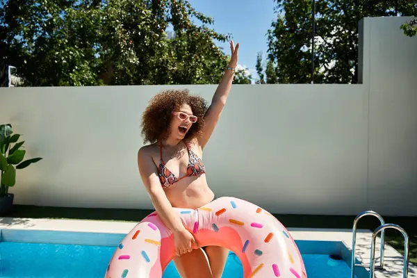 A woman with curly hair, wearing a bikini, enjoys a sunny day by the pool while holding a donut-shaped pool float. — Stock Photo