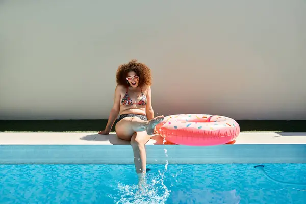 A woman in a bikini enjoys the summer sun by a pool, kicking her feet into the water with a playful smile. — Stock Photo