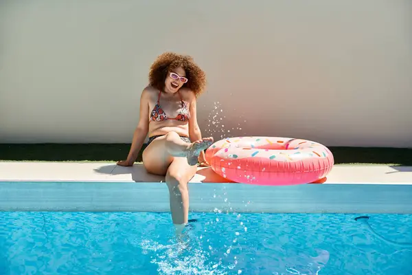 A woman with curly hair in a bikini sits by a pool, dipping her foot into the water while smiling. A pink donut float lies nearby. — Stock Photo