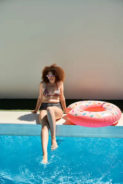 A beautiful woman with curly hair relaxes by the pool in a swimsuit, dipping her toes in the water. — Stock Photo