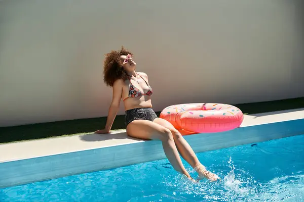 A woman with curly hair enjoys a sunny summer day by the pool, her feet dipping into the cool water. — Stock Photo