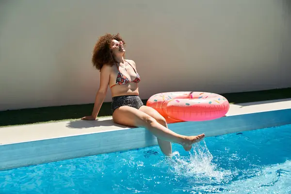 A woman with curly hair in a bikini sits by a pool, dipping her toes in the water while a donut float rests nearby. — Stock Photo