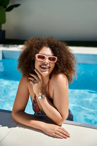 Une femme aux cheveux bouclés et aux lunettes de soleil sourit en s'appuyant contre le bord d'une piscine par une journée ensoleillée. — Photo de stock