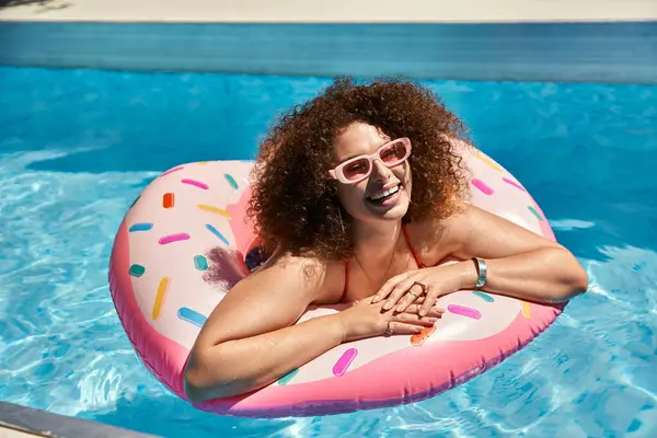 A happy woman with curly hair relaxes on a pink donut float in a swimming pool. — Stock Photo