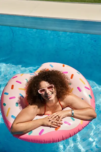 A woman with beautiful curly hair smiles brightly as she floats on a pink donut pool float. — Stock Photo