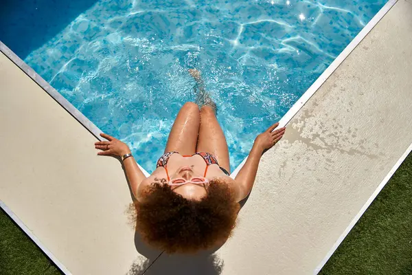 A woman with curly hair sits on the edge of a pool, enjoying the summer sun. — Stock Photo