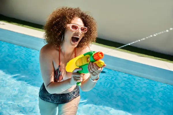 Una donna con i capelli ricci in bikini sorride e spara una pistola ad acqua in piscina in una giornata estiva soleggiata. — Foto stock