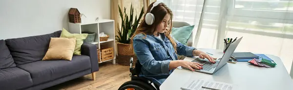 A young woman in a wheelchair sits at a desk in her modern apartment, working on her laptop while wearing headphones. — Stock Photo