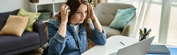 Eine junge Frau im Rollstuhl sitzt an einem Schreibtisch in einer modernen Wohnung und benutzt Laptop und Kopfhörer. — Stockfoto