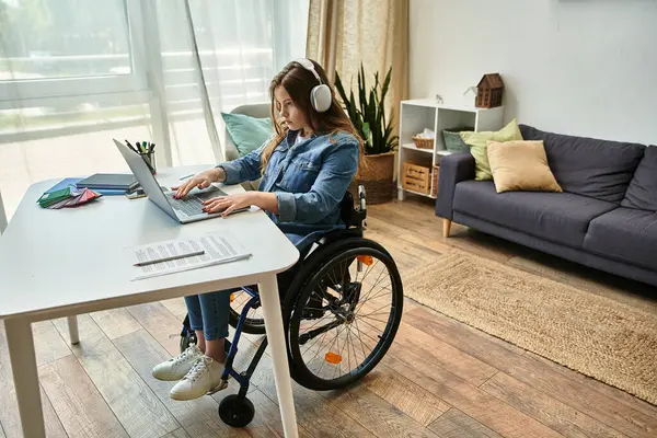 Uma jovem mulher em uma cadeira de rodas senta-se em sua mesa, trabalhando em um laptop, fones de ouvido em, em seu apartamento moderno. — Fotografia de Stock