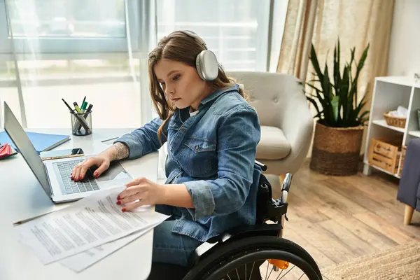 A young woman in a wheelchair works on a laptop while wearing headphones in her modern apartment. — Stock Photo