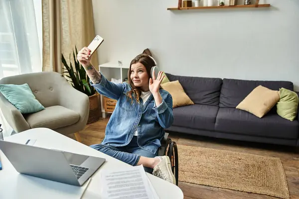 A young woman in a wheelchair is taking a selfie in her modern apartment. She is wearing headphones and smiling at the camera. — Stock Photo