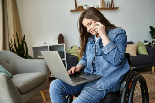 A young woman in a wheelchair uses her laptop and phone, sitting in a contemporary apartment living room. — Stock Photo
