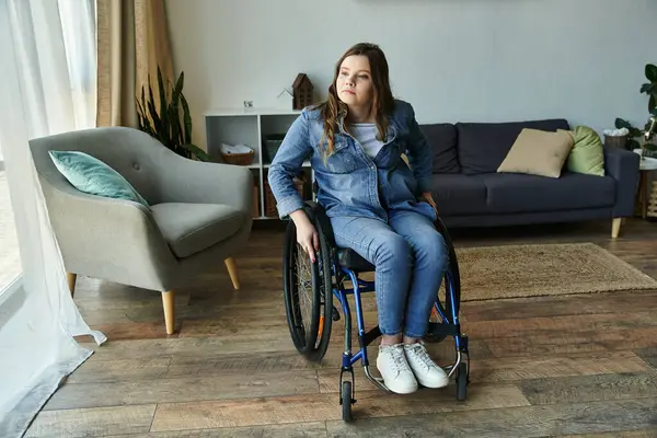 A young woman in a wheelchair sits in a modern apartment, with a large window and a couch in the background. — Stock Photo