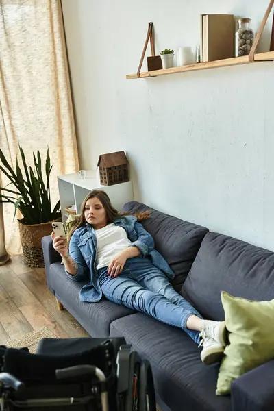 A young woman in a wheelchair relaxes on a comfortable couch in her modern apartment, using her phone. — Stock Photo