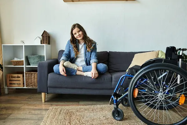 A young woman with long brown hair sits on a gray couch in a modern apartment, her wheelchair positioned nearby. — Stock Photo