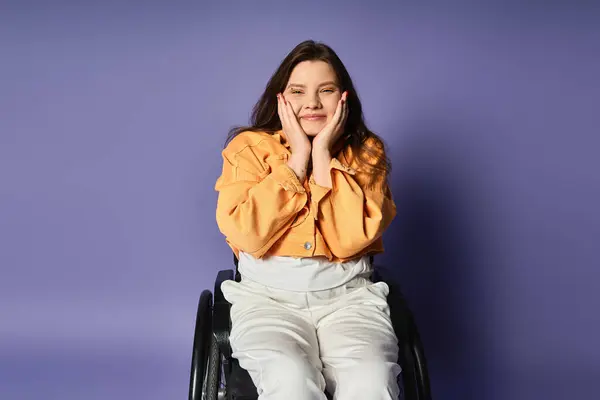 A young woman in casual clothing sits in a wheelchair against a purple backdrop. She smiles with her hands on her cheeks. — Stock Photo