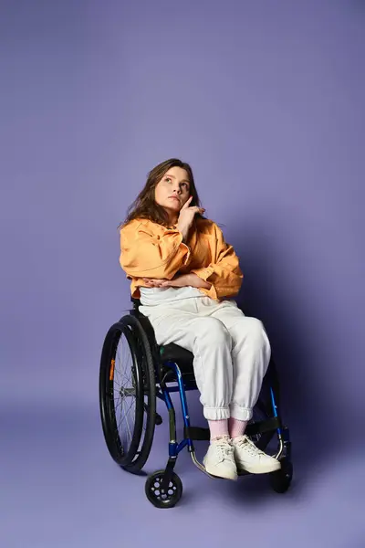 Uma mulher em uma cadeira de rodas, vestida com uma camisa amarela e calças brancas, contempla em um estúdio roxo. — Fotografia de Stock