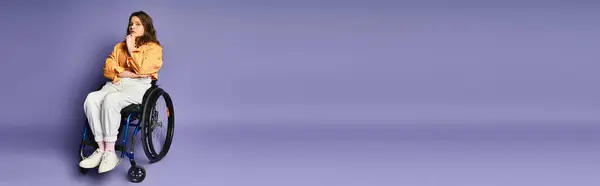 A young woman in casual attire sits thoughtfully in a wheelchair against a purple background. — Stock Photo