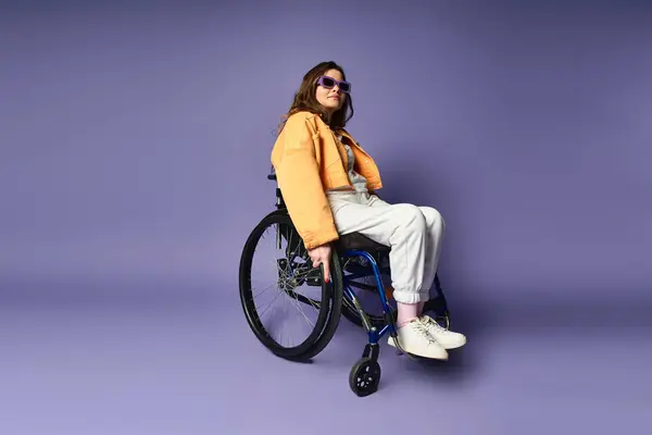 A young woman in trendy casual clothing sits in a wheelchair in a studio setting with a purple background. — Stock Photo