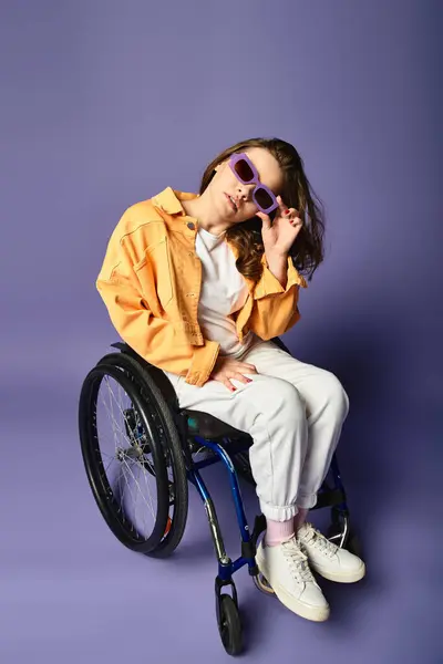 Young woman in trendy casual attire and wheelchair sits in studio with purple background, holding sunglasses up to face, facing camera — Stock Photo