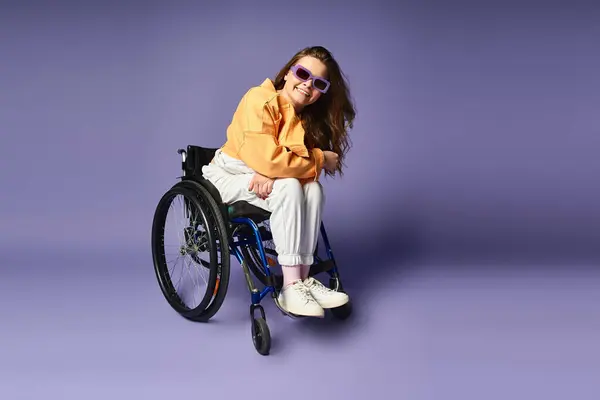 Young woman in wheelchair, wearing trendy clothes and sunglasses, smiling at camera in studio with purple background — Stock Photo